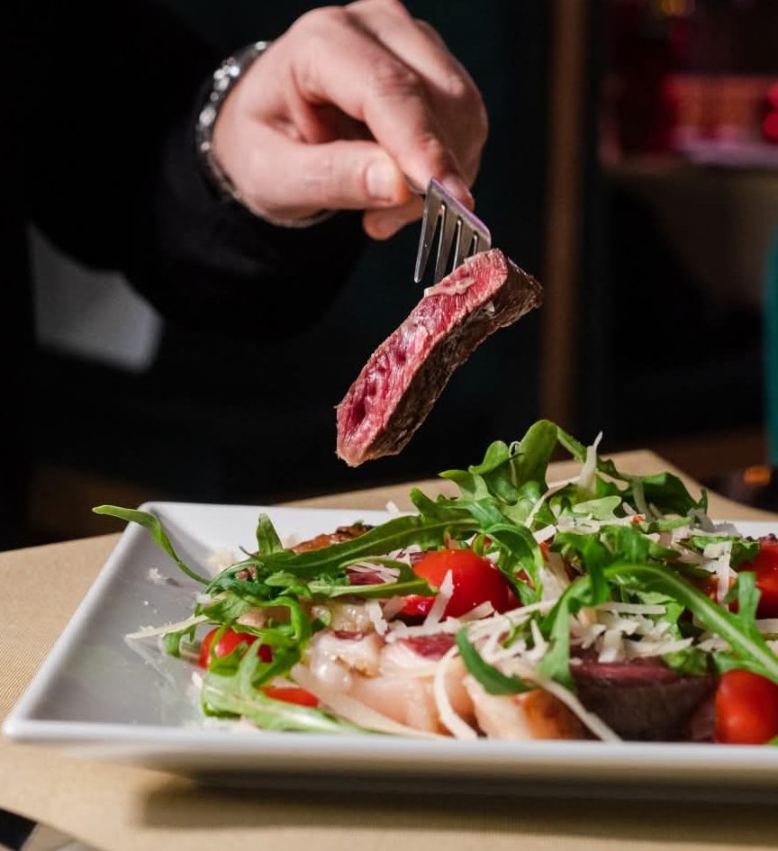 Fetta di tagliata rosata sollevata con la forchetta sopra rucola, pomodorini e grana